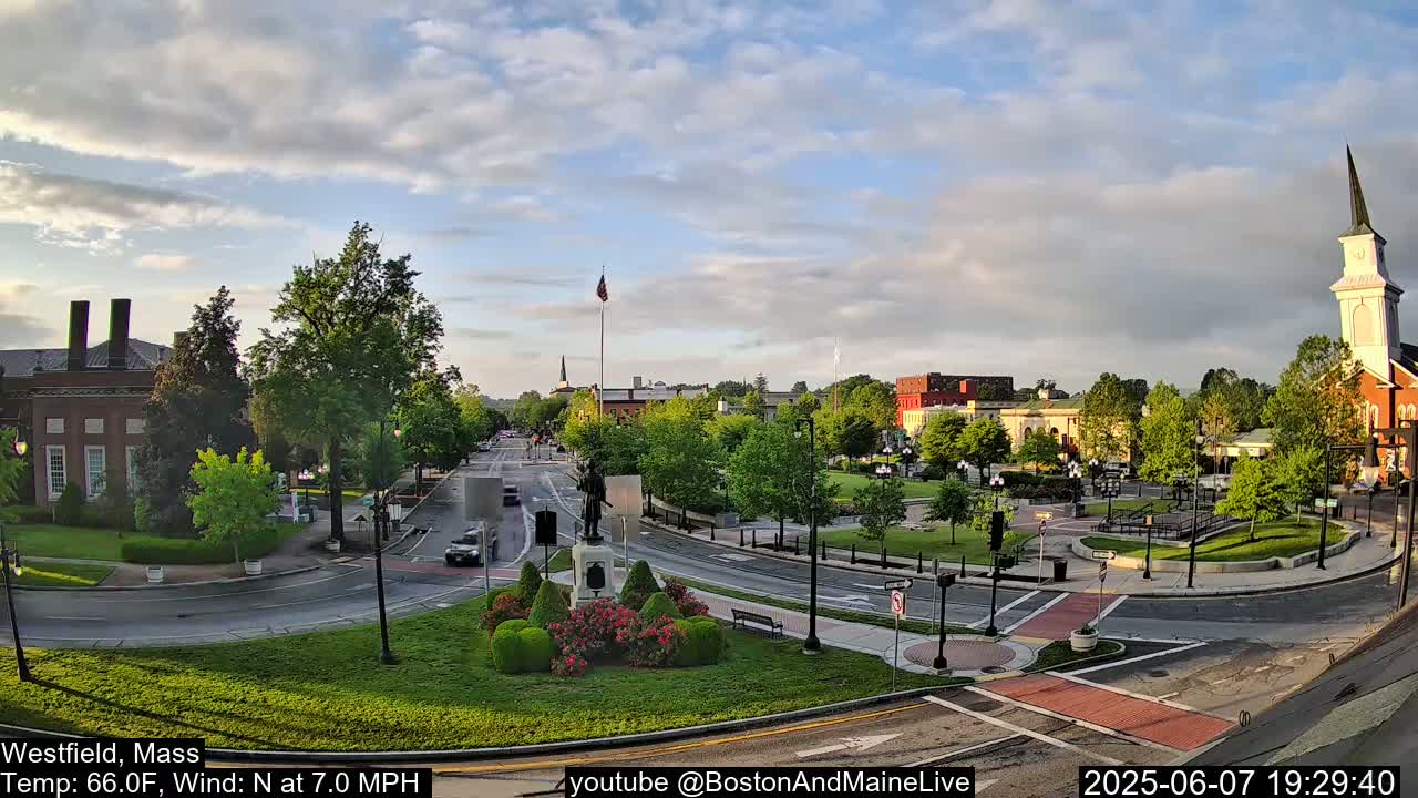 A partly cloudy evening scene shows a town square with a statue, surrounding buildings, and a church steeple, with cars on the roads.
