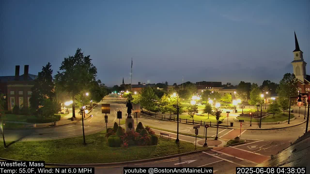 A town square at twilight, illuminated by streetlights, with a statue in the center and a church steeple in the background under a clear, dark sky.
