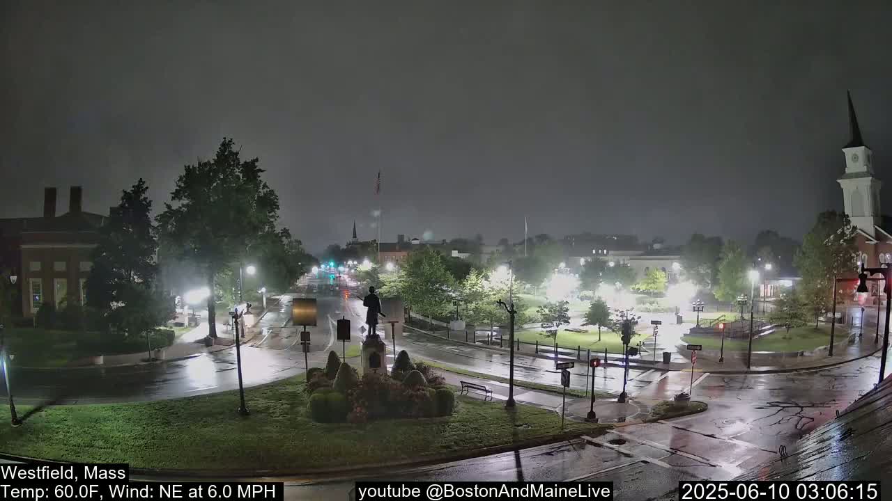 A nighttime, rainy scene shows a town square with a statue in the center, streetlights illuminating wet roads, and buildings in the background.