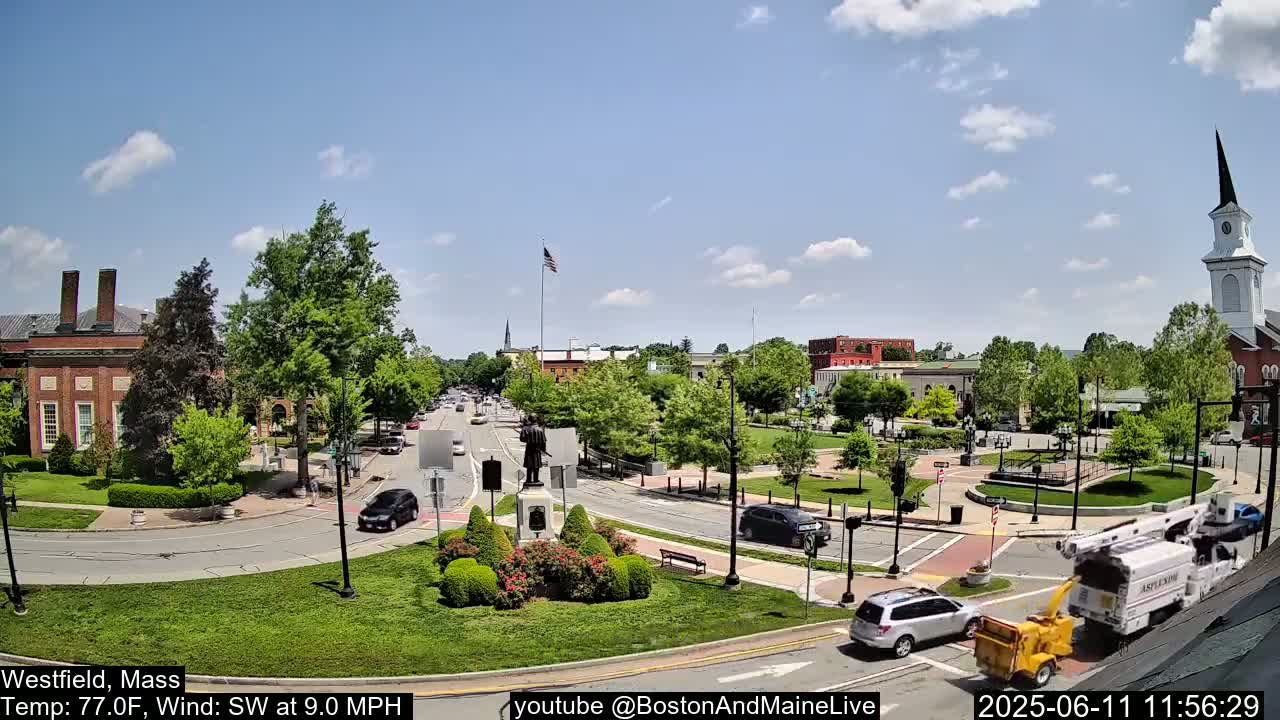 A town square with a statue at its center, surrounded by brick buildings, lush green trees, and cars under a partly cloudy sky.