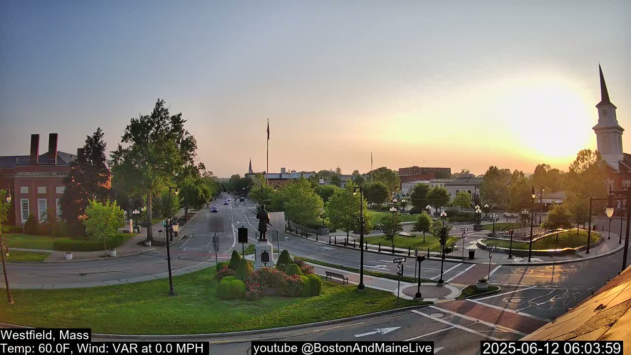 A town square at sunrise, with calm weather, features a statue in a landscaped roundabout, surrounded by streets, buildings, and trees.