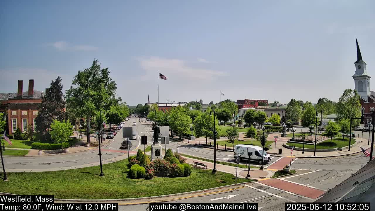 A sunny town square with a statue in the center, surrounded by brick buildings, green spaces, and roadways, under a mostly clear sky.
