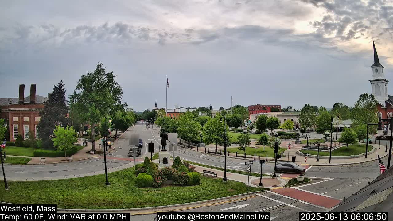 A mostly cloudy sky hangs over a town square with a statue, green spaces, brick buildings, and a church steeple.