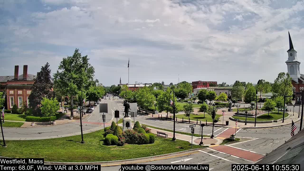 A partly cloudy day reveals a town square with a statue in the center, surrounded by buildings, trees, and a church steeple.