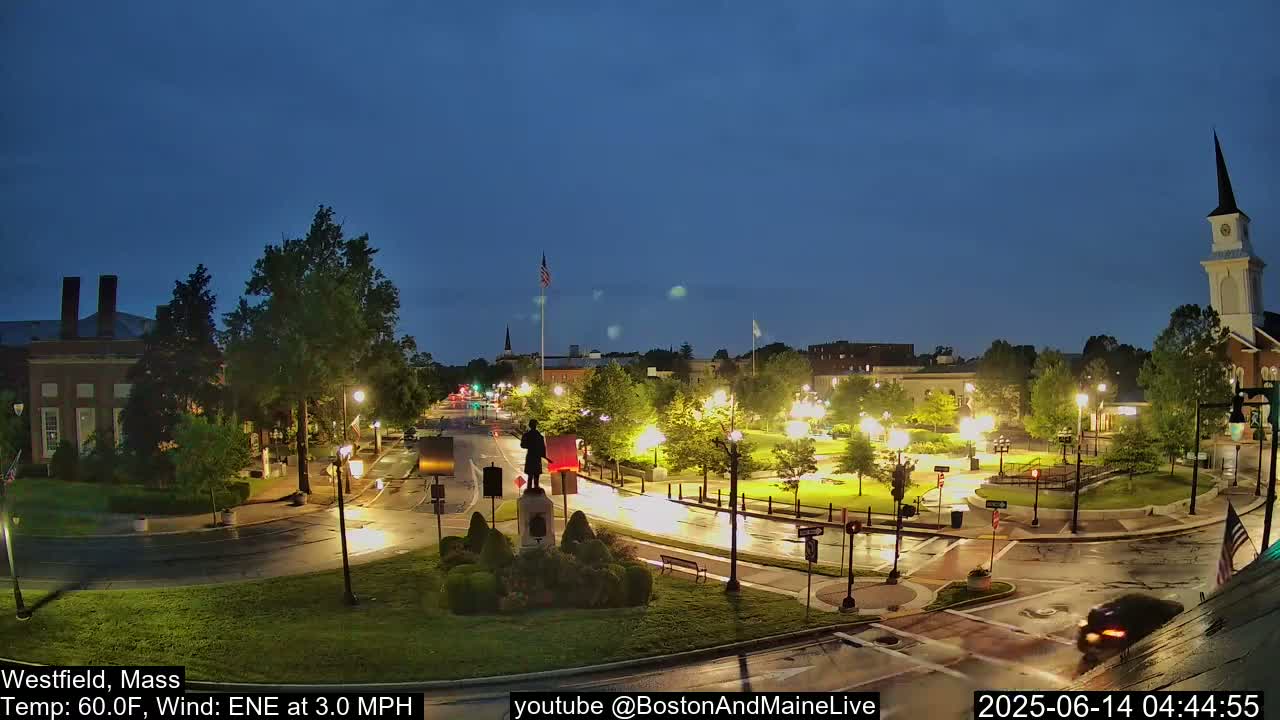 A nighttime view of a town square with a statue at its center, illuminated by streetlights under a twilight sky.