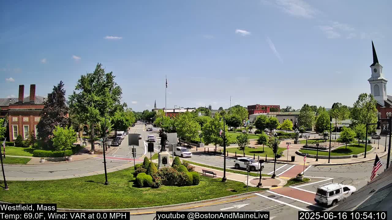 A sunny town square with a monument in the center, surrounded by brick buildings, green spaces, and cars on the road.