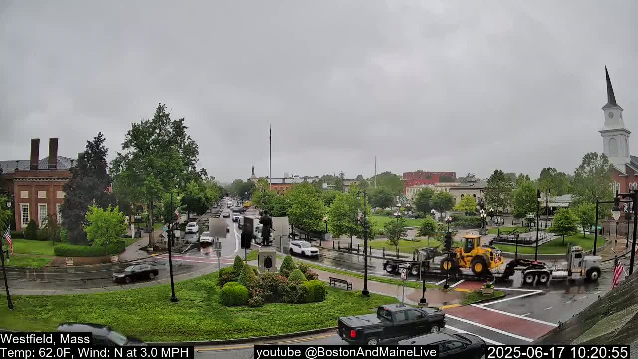 A rainy town square with several cars and trucks at an intersection, under a gray sky.
