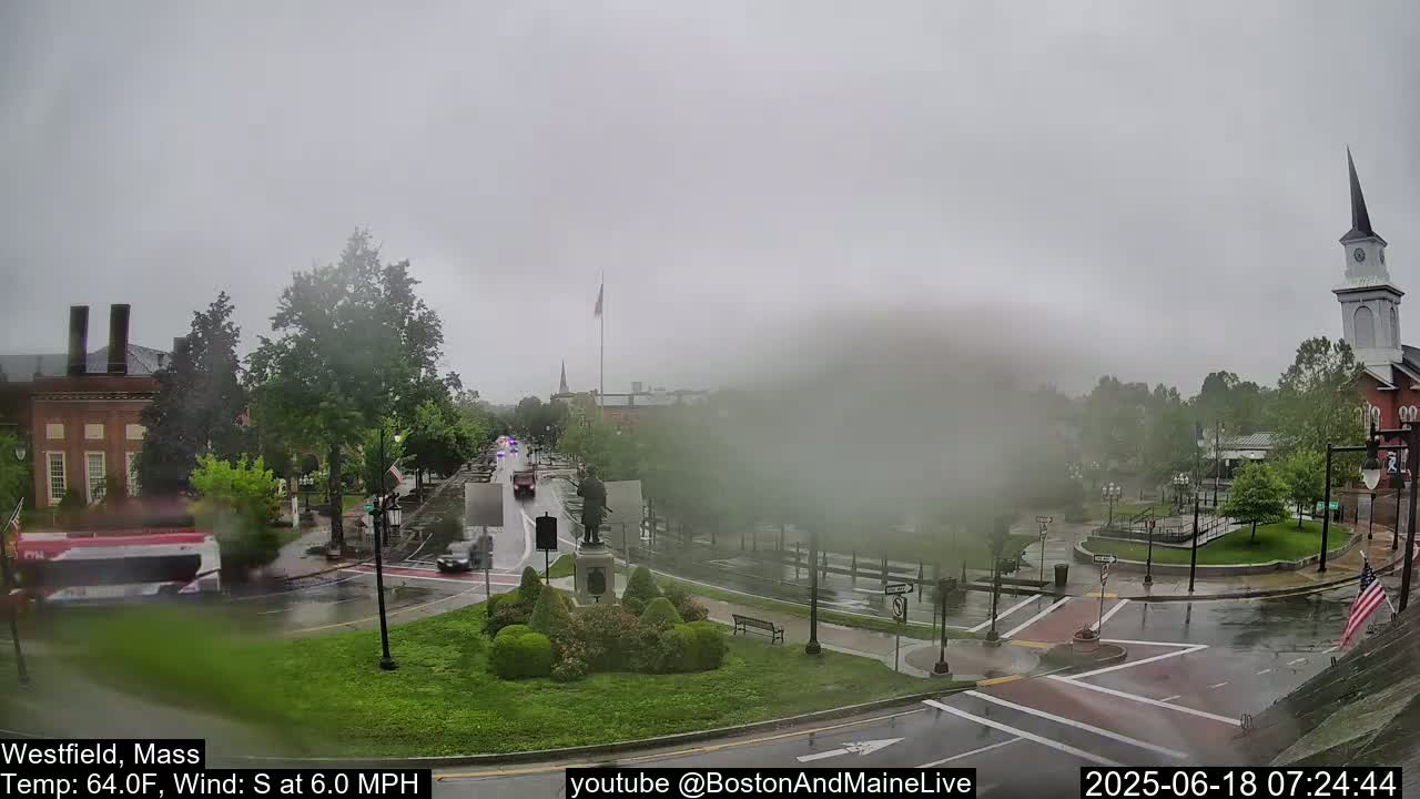 A town square with several buildings and a church steeple is seen on a rainy day with light traffic.