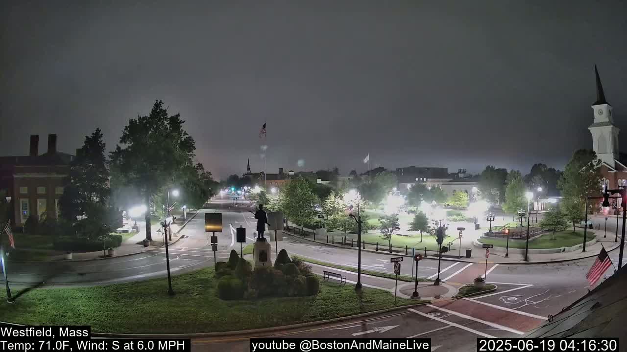 A nighttime view of a town square with a statue, a church steeple, and several buildings under a hazy, overcast sky.
