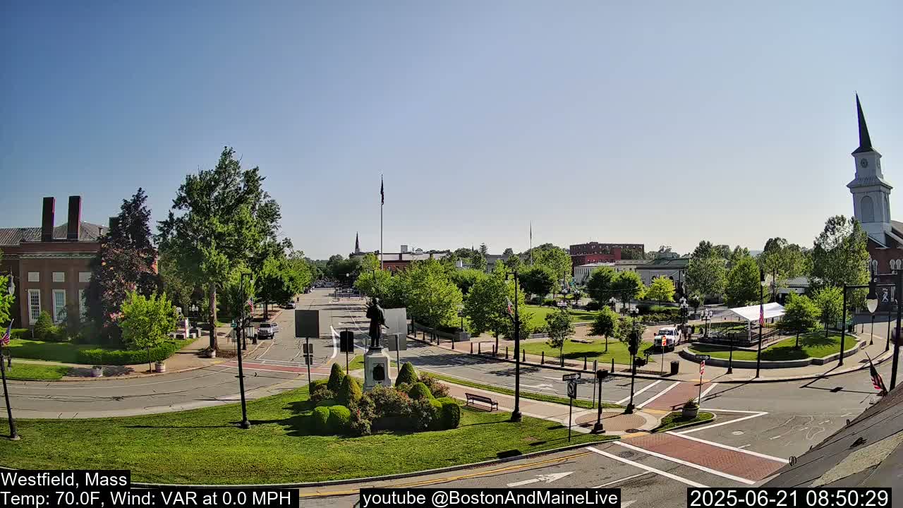 A town square with a statue, surrounded by green trees and brick buildings, on a sunny day.