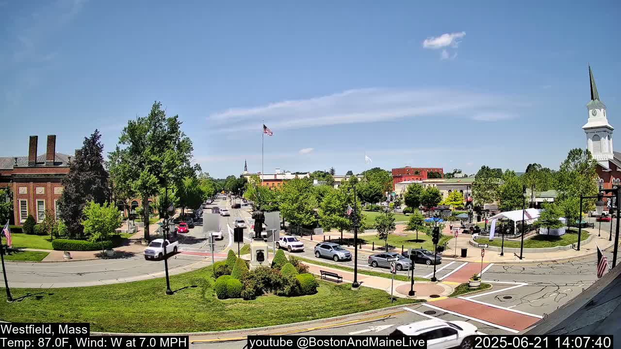 A town square with a monument at its center, surrounded by brick buildings, green spaces, and cars on a sunny day with a light wind.
