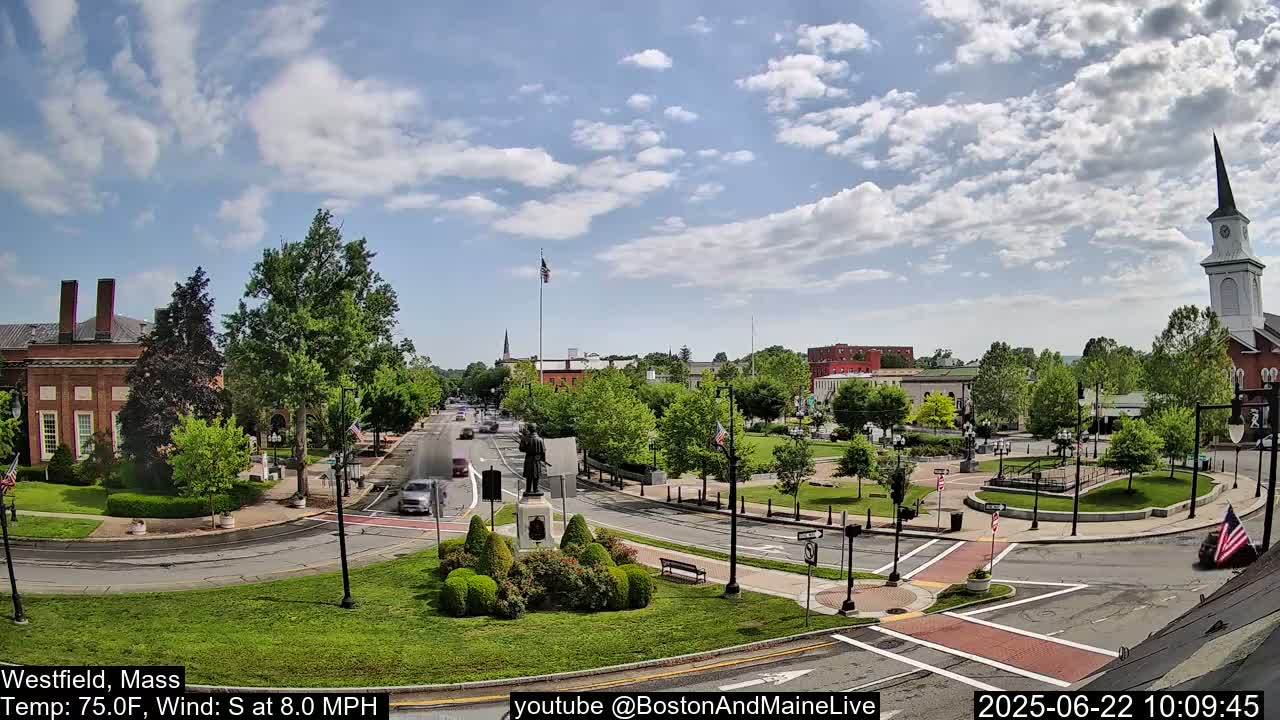 A partly sunny day shows a town square with a monument, green spaces, brick buildings, and a church steeple.