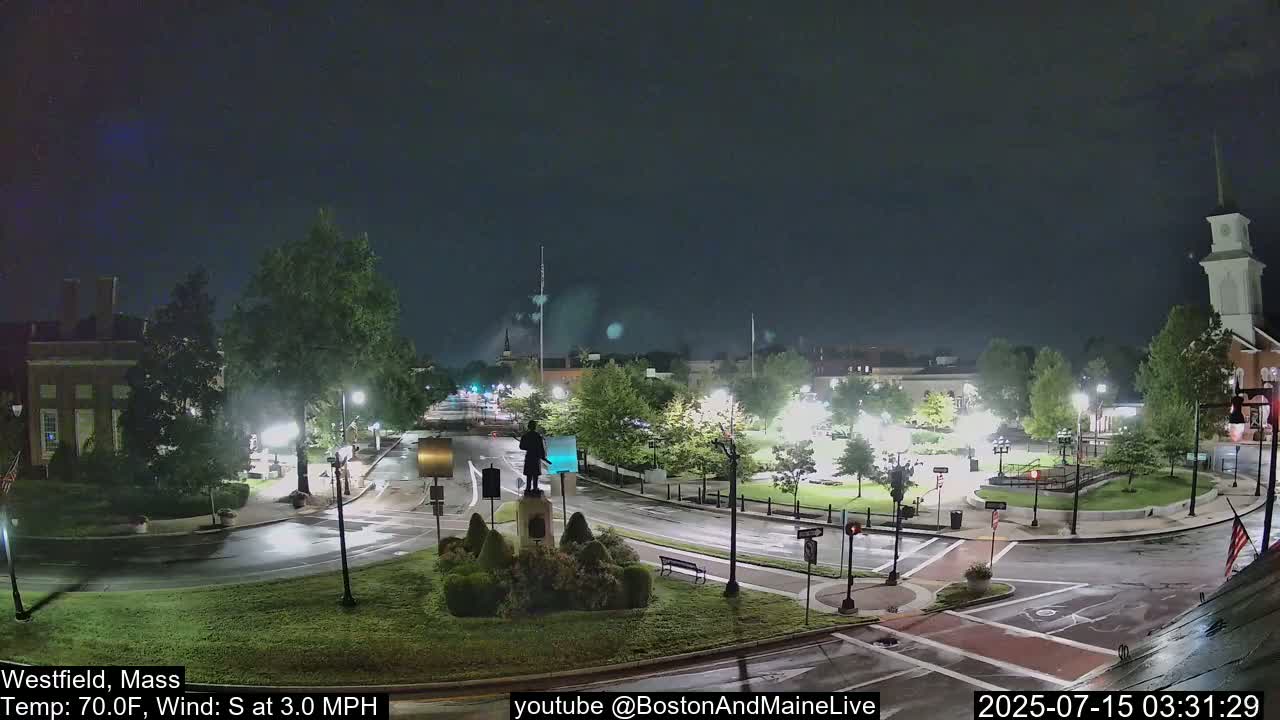A nighttime view of a town square with a statue in the center, light rain falling, and a few cars on the roads.