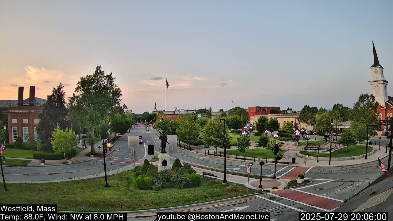 A nighttime view of a town square with a statue in the center, light rain falling, and a few cars on the roads.