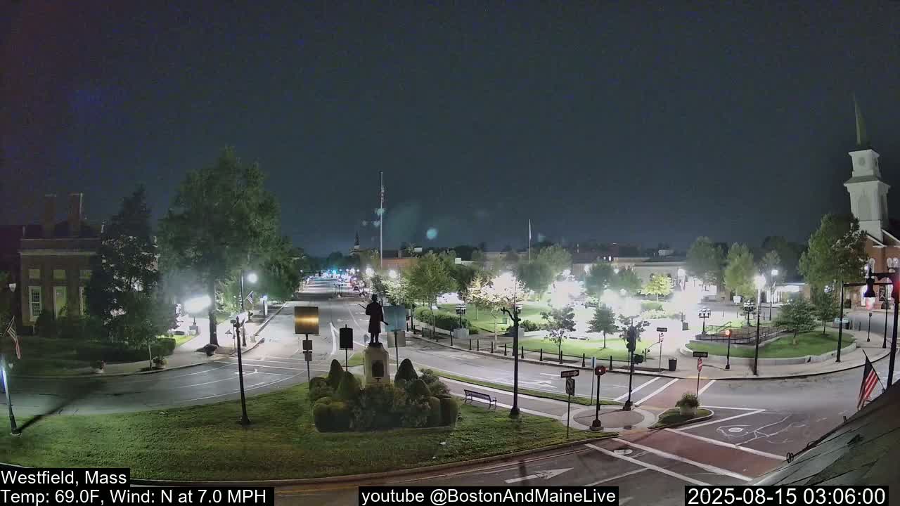 A nighttime view of a town square with a statue in the center, under a clear, dark sky with a 7 mph north wind and a temperature of 69 degrees Fahrenheit.