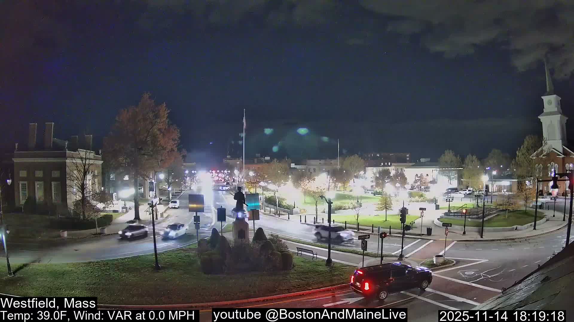 A nighttime view captures a busy town square featuring a central rotary with a statue, a historical building, a prominent white-steepled church, and multiple cars in motion, all illuminated by streetlights under a cloudy, cold, and calm sky.