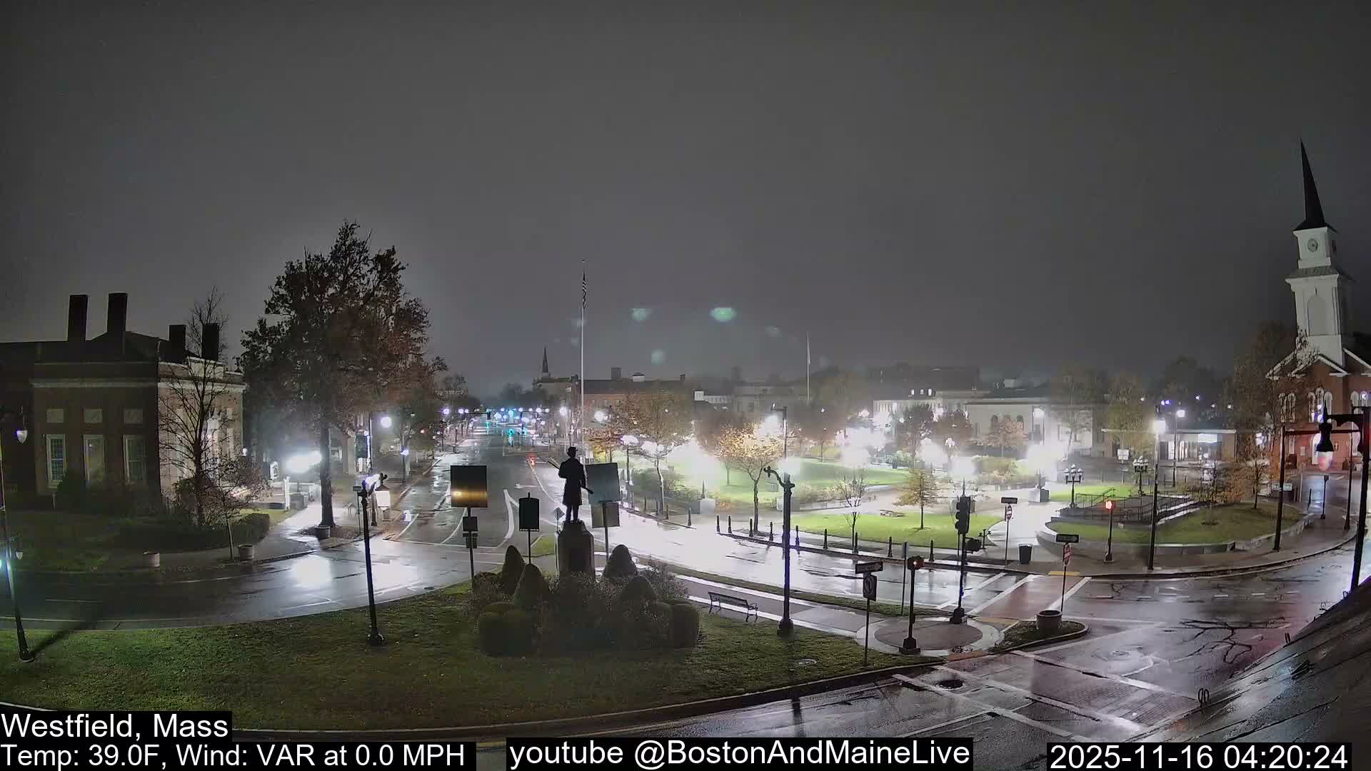 The image presents an illuminated town square at night or early morning, with wet streets and sidewalks reflecting the numerous streetlights and building lights under a dark, overcast sky, hinting at recent rain.