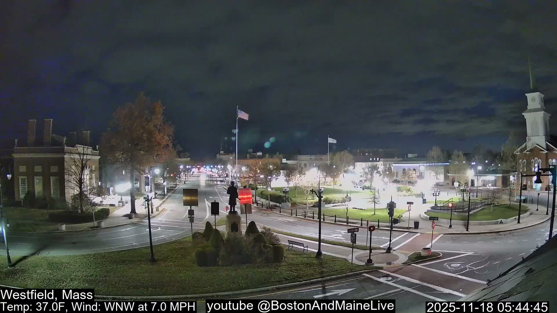 The image presents an illuminated town square at night or early morning, with wet streets and sidewalks reflecting the numerous streetlights and building lights under a dark, overcast sky, hinting at recent rain.