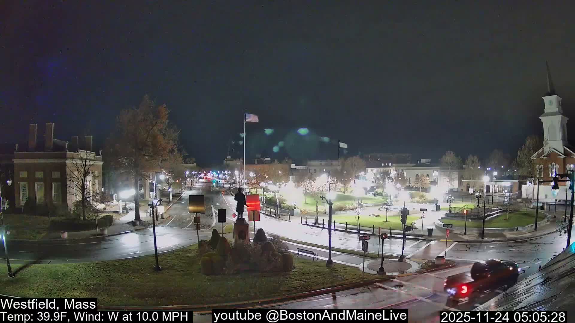 The image displays a dark, wet town square at night, illuminated by numerous streetlights, with a central statue, various buildings including a church with a steeple, and a car moving on the glistening roads.