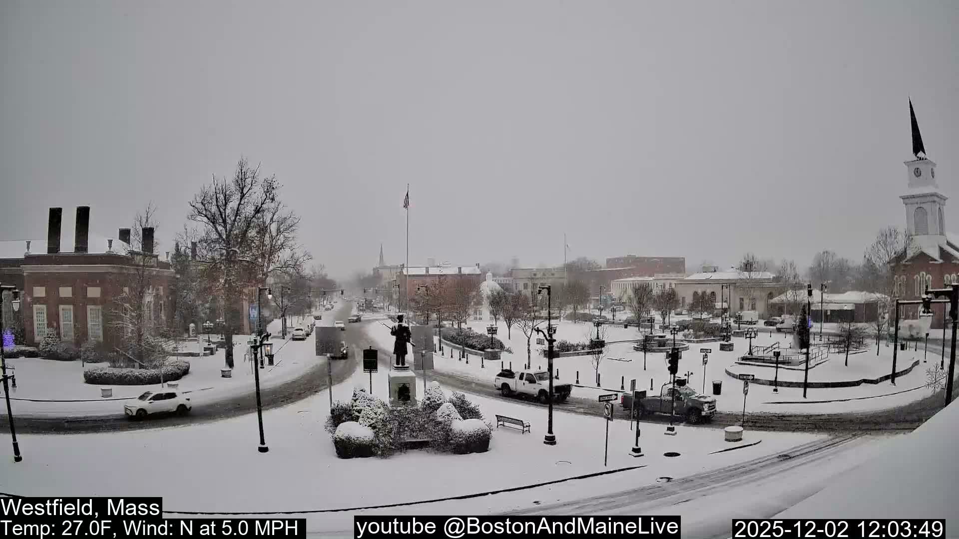 A town square and its surrounding buildings are covered in fresh snow under an overcast sky, with vehicles on the roads and a statue in the central island.