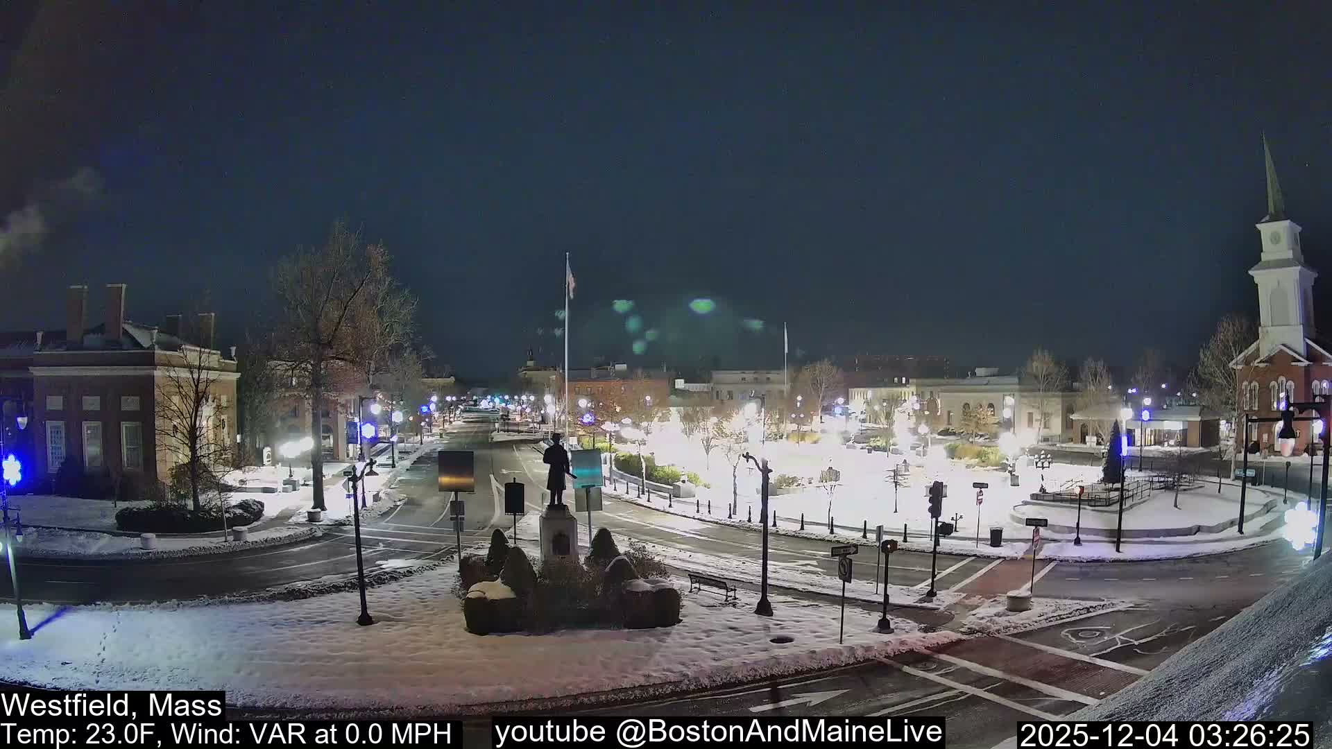 A snow-dusted town square and intersection are brightly illuminated by streetlights and building lights on a clear, cold night.