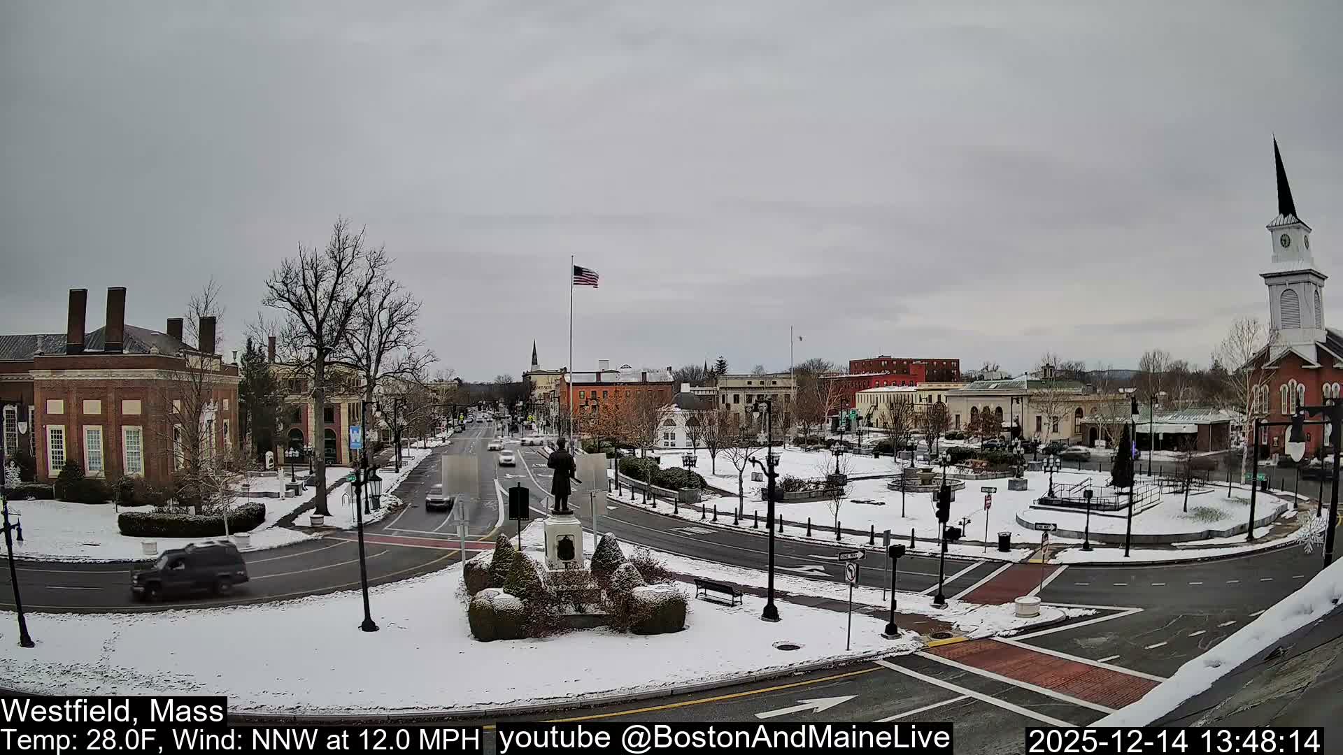 A snow-dusted town square and intersection are brightly illuminated by streetlights and building lights on a clear, cold night.