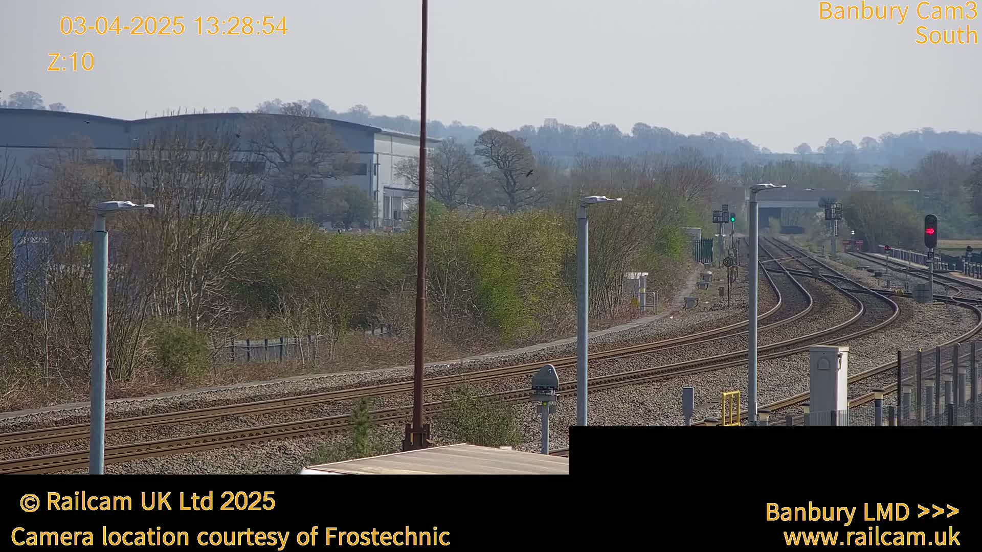 A sunny view of multiple train tracks curving through a landscape of trees, shrubs, and industrial buildings.