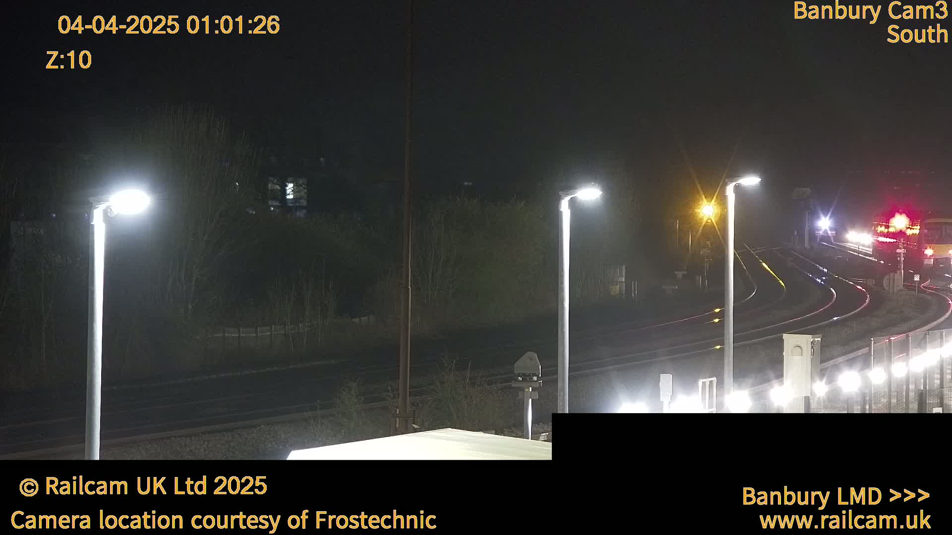 A nighttime view of a train station with several lights illuminating railway tracks and a train in the distance.