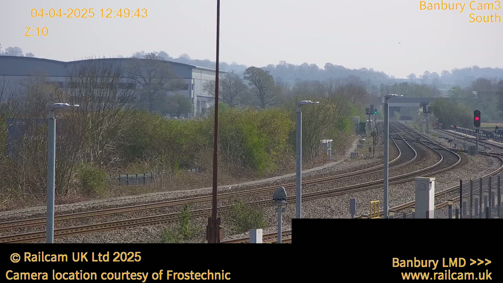 A sunny view of a railway track branching off into several directions, with a large industrial building and trees in the background.