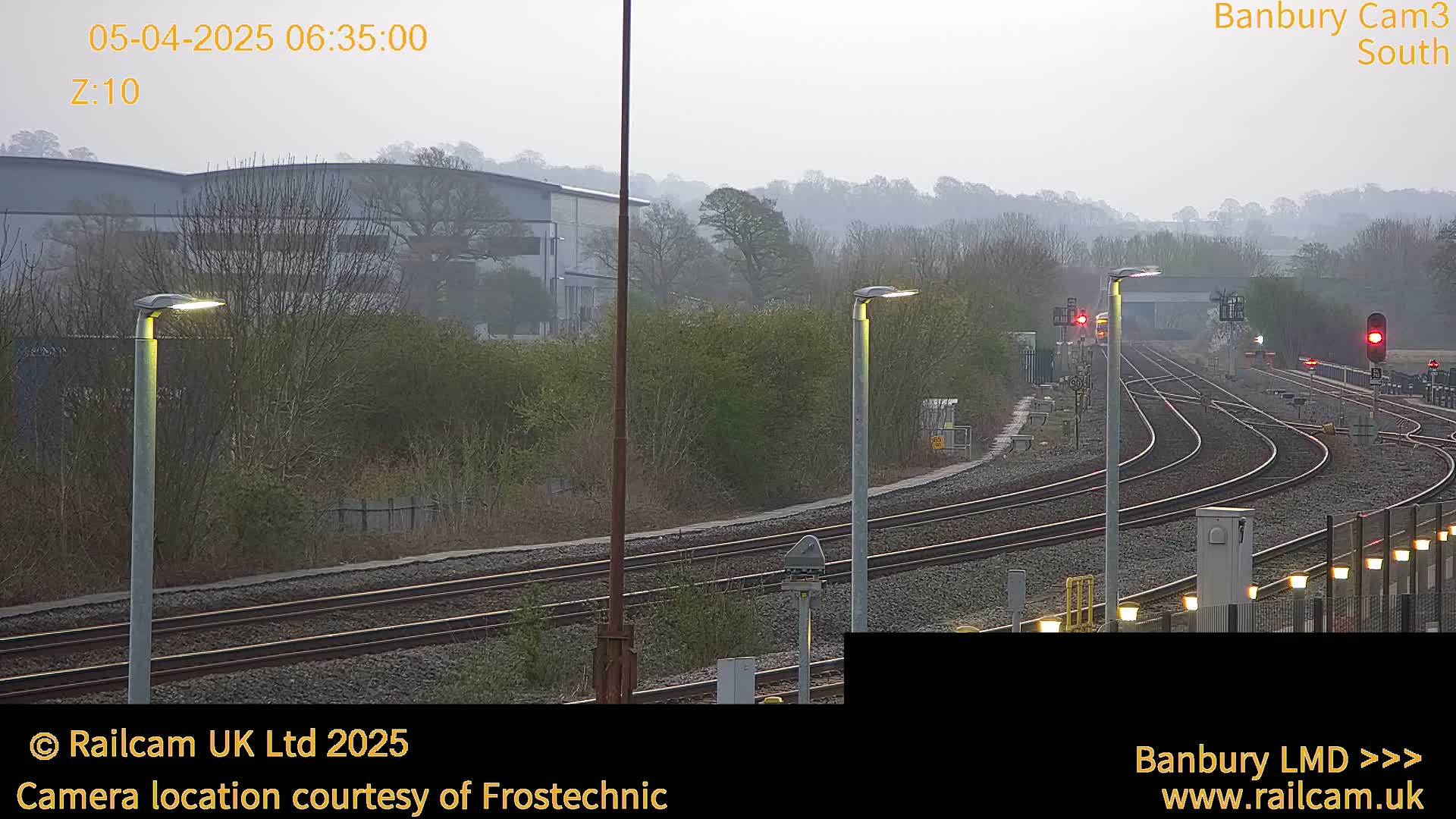 A hazy view of a railway track system curving gently away from the camera, past several lampposts and a building in the distance.