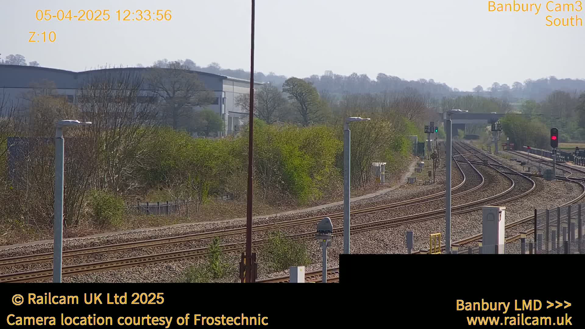A sunny outdoor view of a railway yard with multiple sets of tracks curving around a small building, bordered by trees and shrubs.