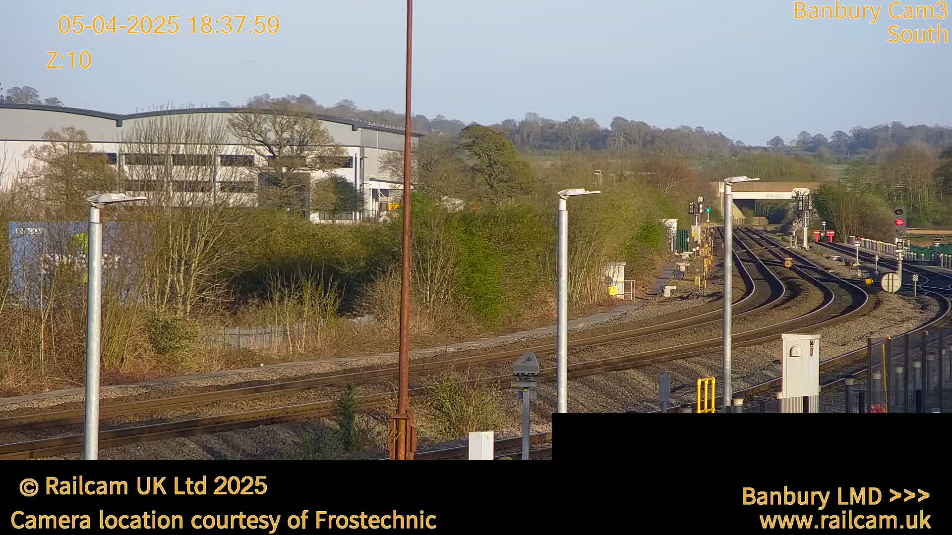 A view of curving train tracks under a bright, clear sky, with industrial buildings and trees in the background.