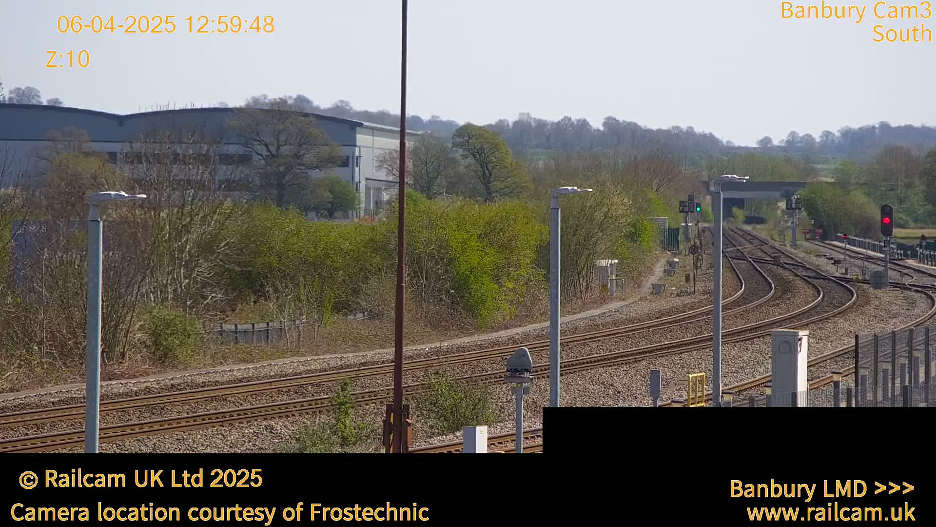 A sunny day view of multiple train tracks curving through a landscape with a warehouse and trees in the background.