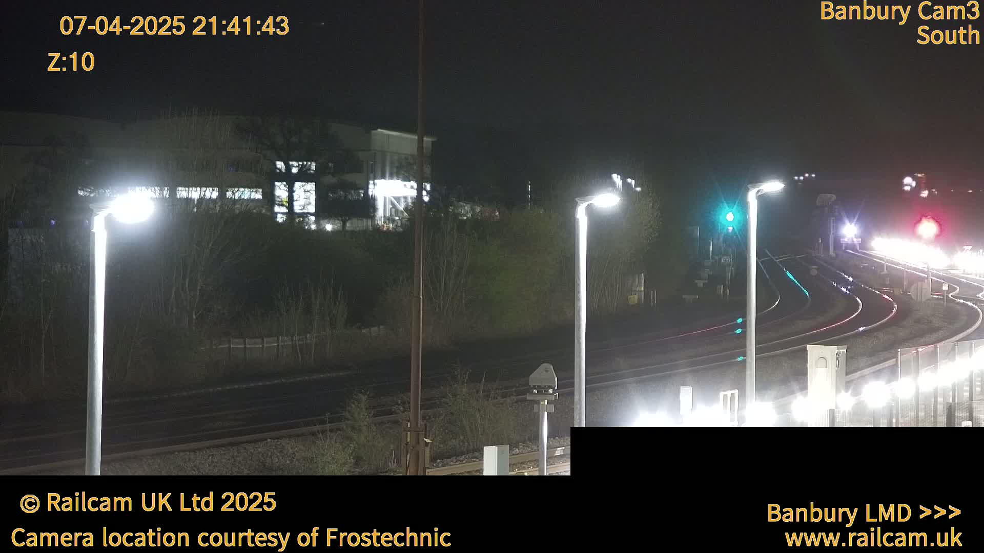 At night, illuminated train tracks curve through a landscape with streetlights and a building in the background.