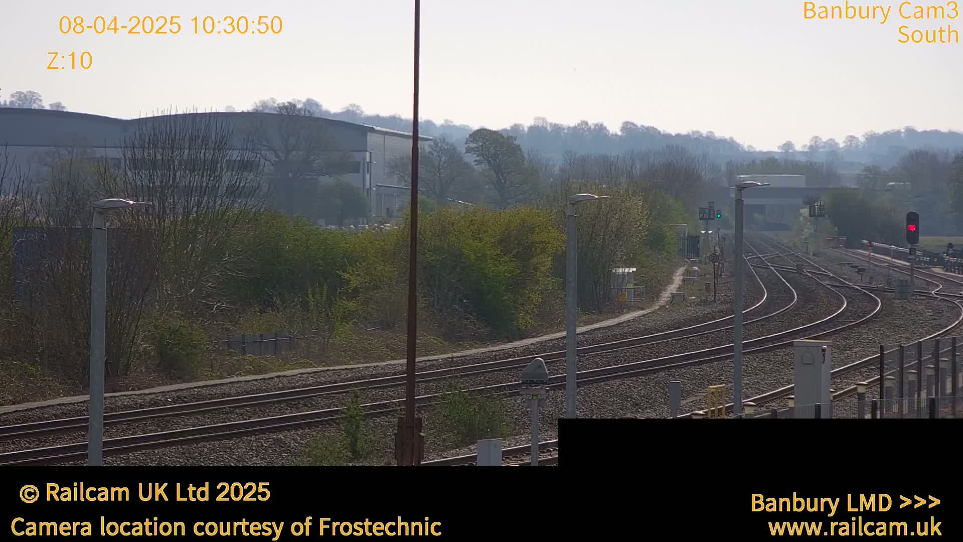A slightly hazy sunny day reveals a view of train tracks curving through a landscape of low-lying vegetation and industrial buildings.