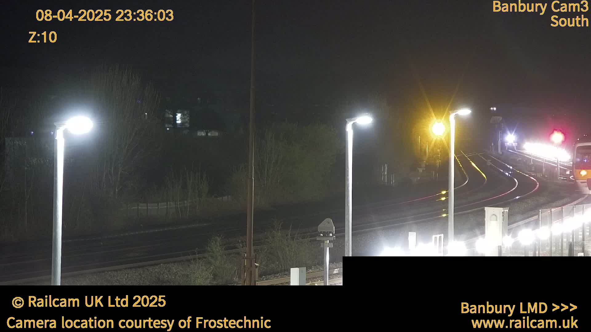 A nighttime, slightly wet view of train tracks and a departing train, illuminated by streetlights.