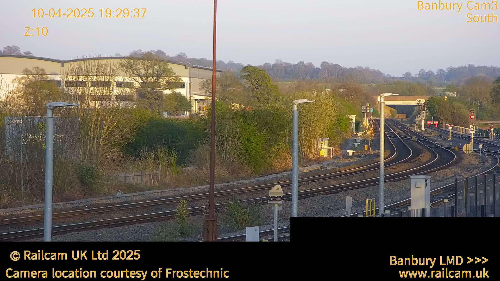 A long shot of a railway track curving through a landscape under a pale evening sky, with buildings and trees in the background.