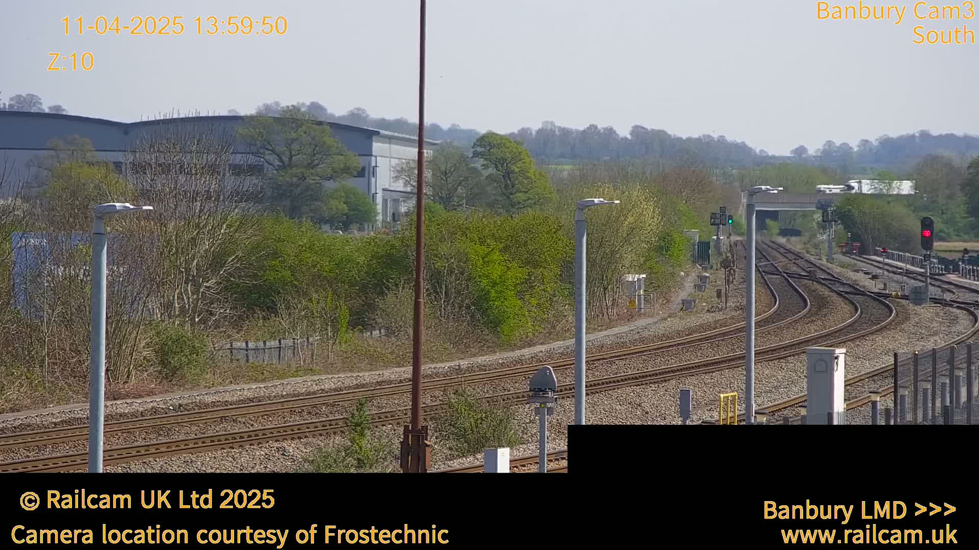 A sunny view of a railway line curving through a landscape of trees, shrubs, and industrial buildings.