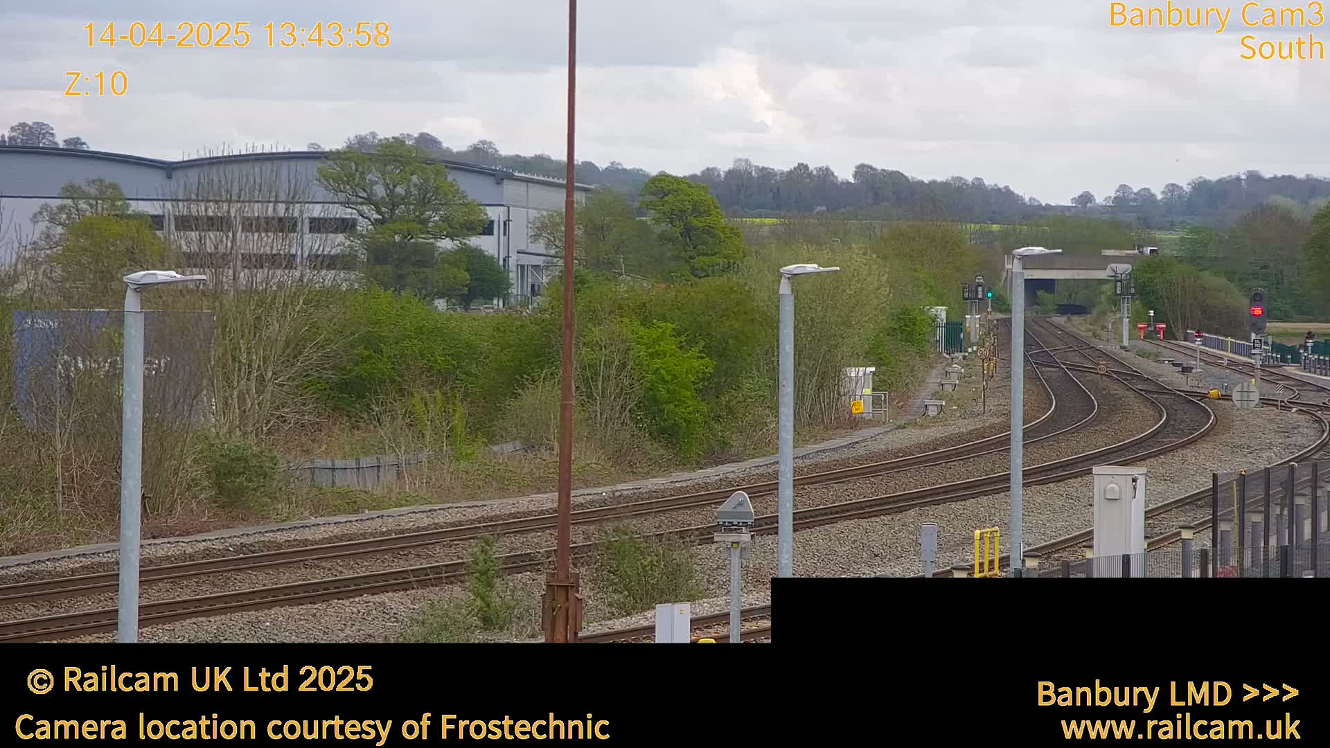 Multiple train tracks curve through a landscape of green vegetation and industrial buildings under an overcast sky.