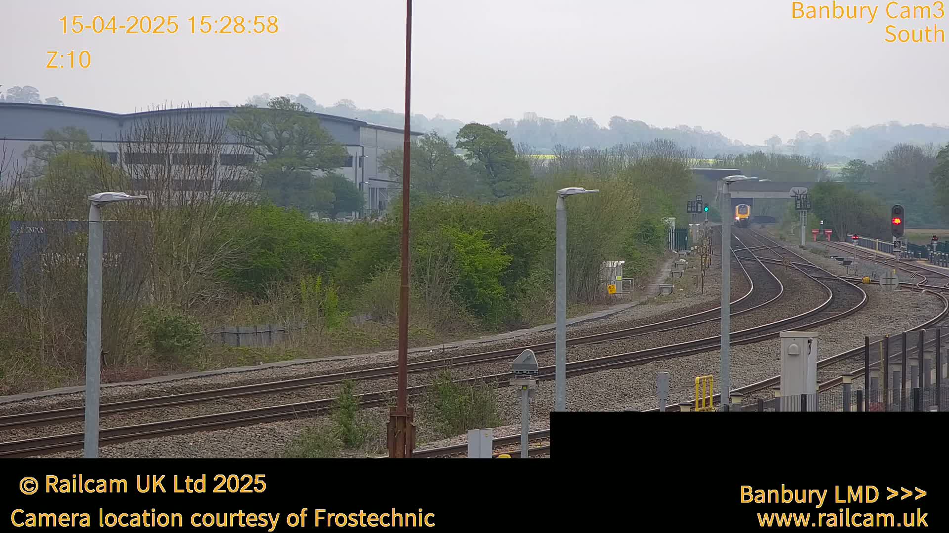 A train approaches a railway station on a hazy day, passing through a curve in the tracks surrounded by trees and industrial buildings.
