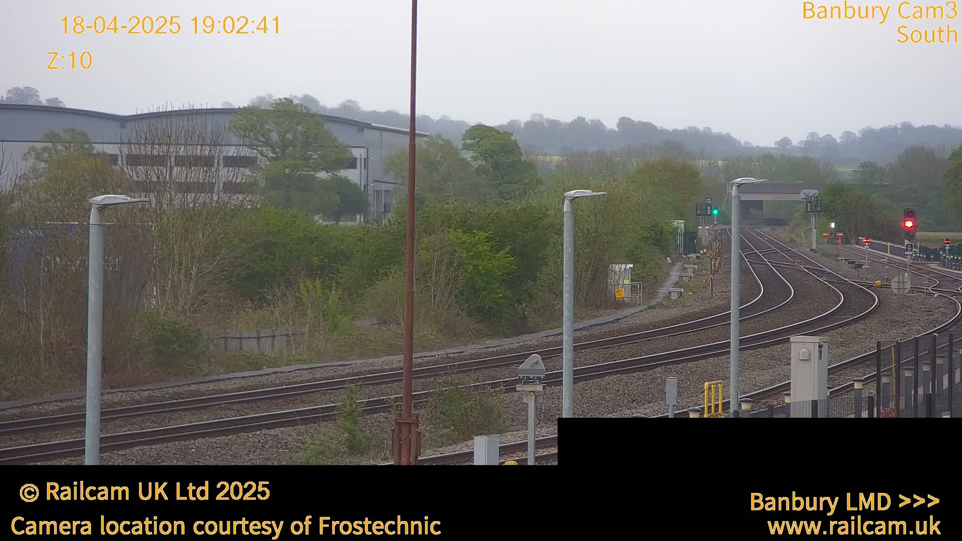 A view of a railway yard with multiple tracks curving away from the camera, under an overcast sky, with industrial buildings and trees in the background.