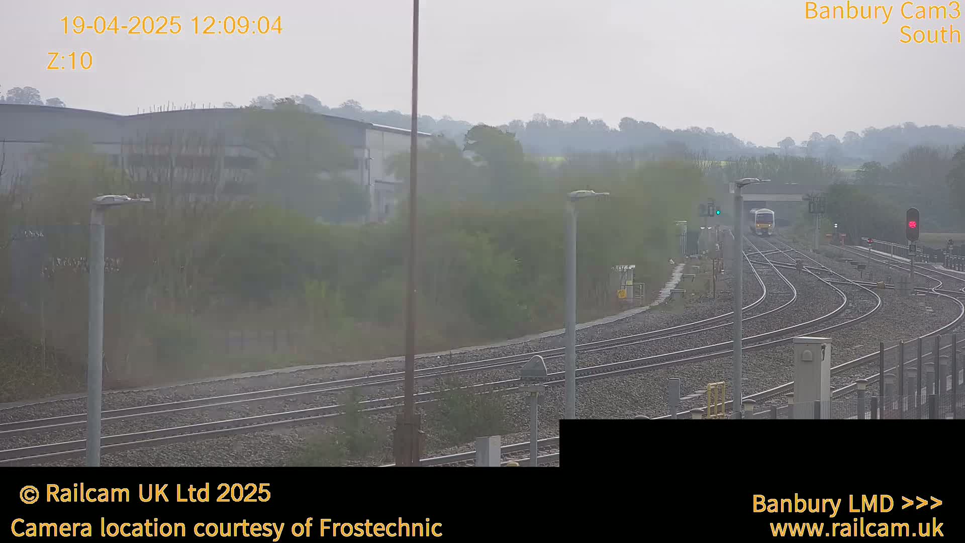 A train approaches a railway junction on an overcast day, passing industrial buildings and greenery.
