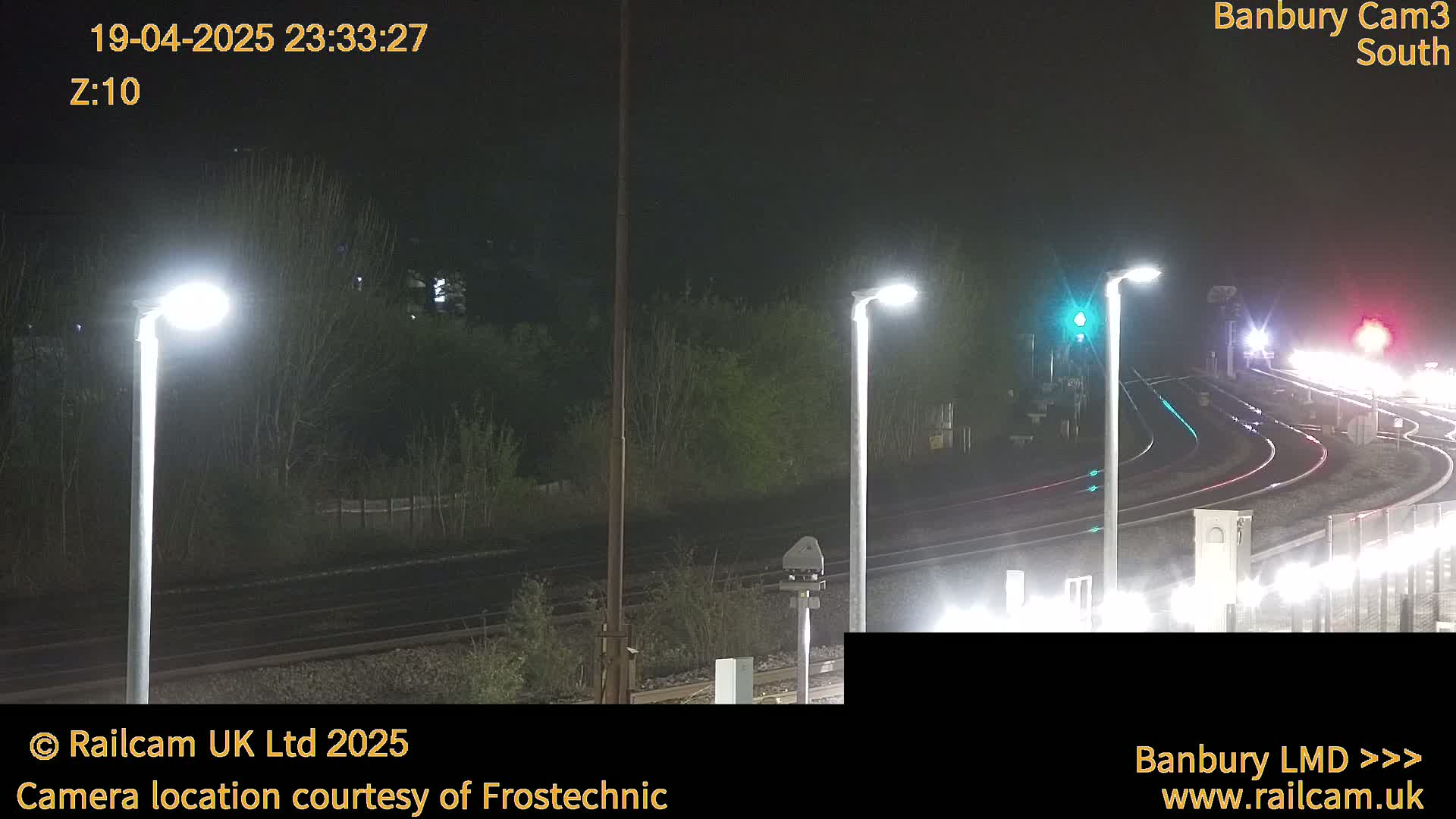 A nighttime view of a train track intersection with several illuminated lampposts, surrounded by dark trees and bushes.