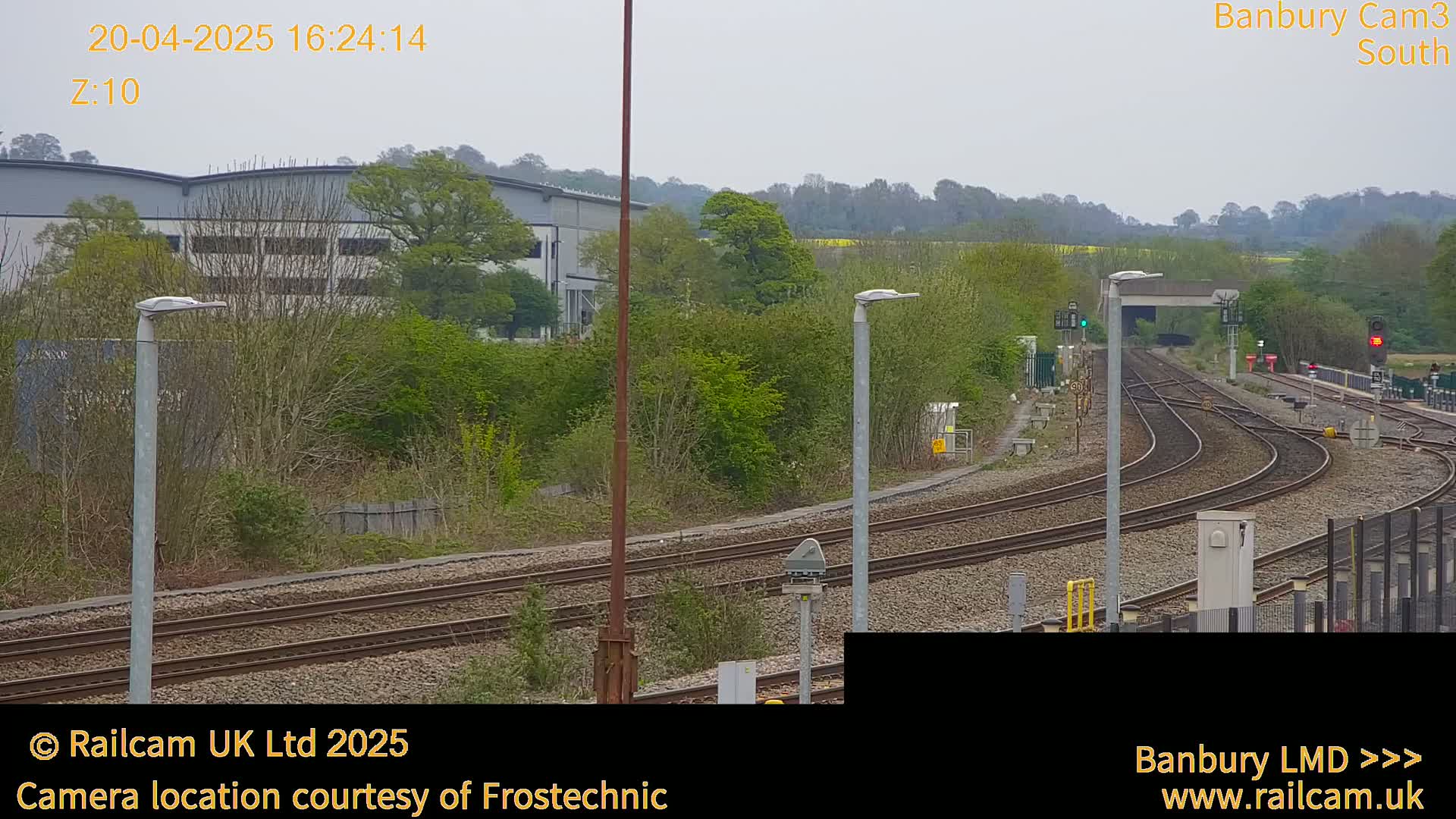A view of several curving train tracks on an overcast day, alongside industrial buildings and green vegetation.