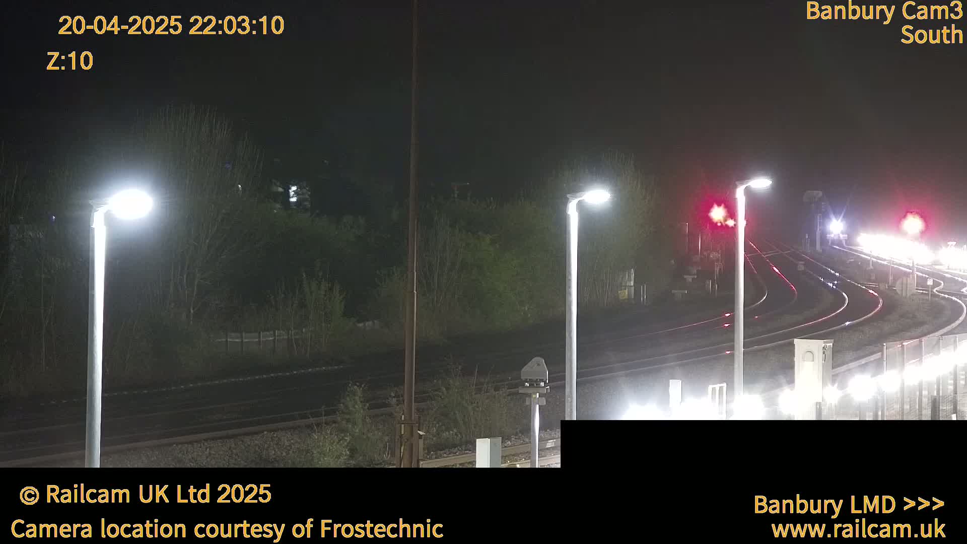 At night, illuminated train tracks curve through a dark landscape under several bright streetlights.