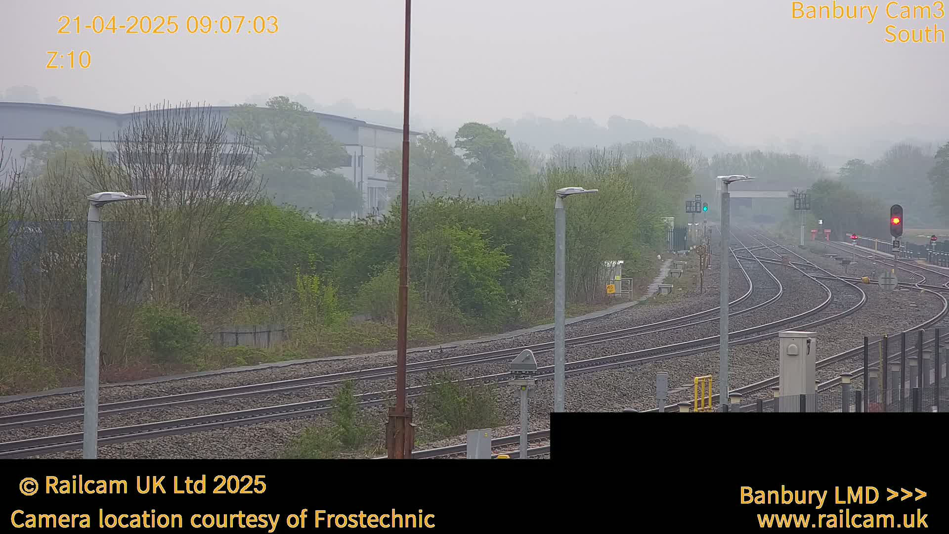 A foggy view of a railway junction with several tracks curving away from the viewer, alongside a hedgerow and industrial buildings in the distance.