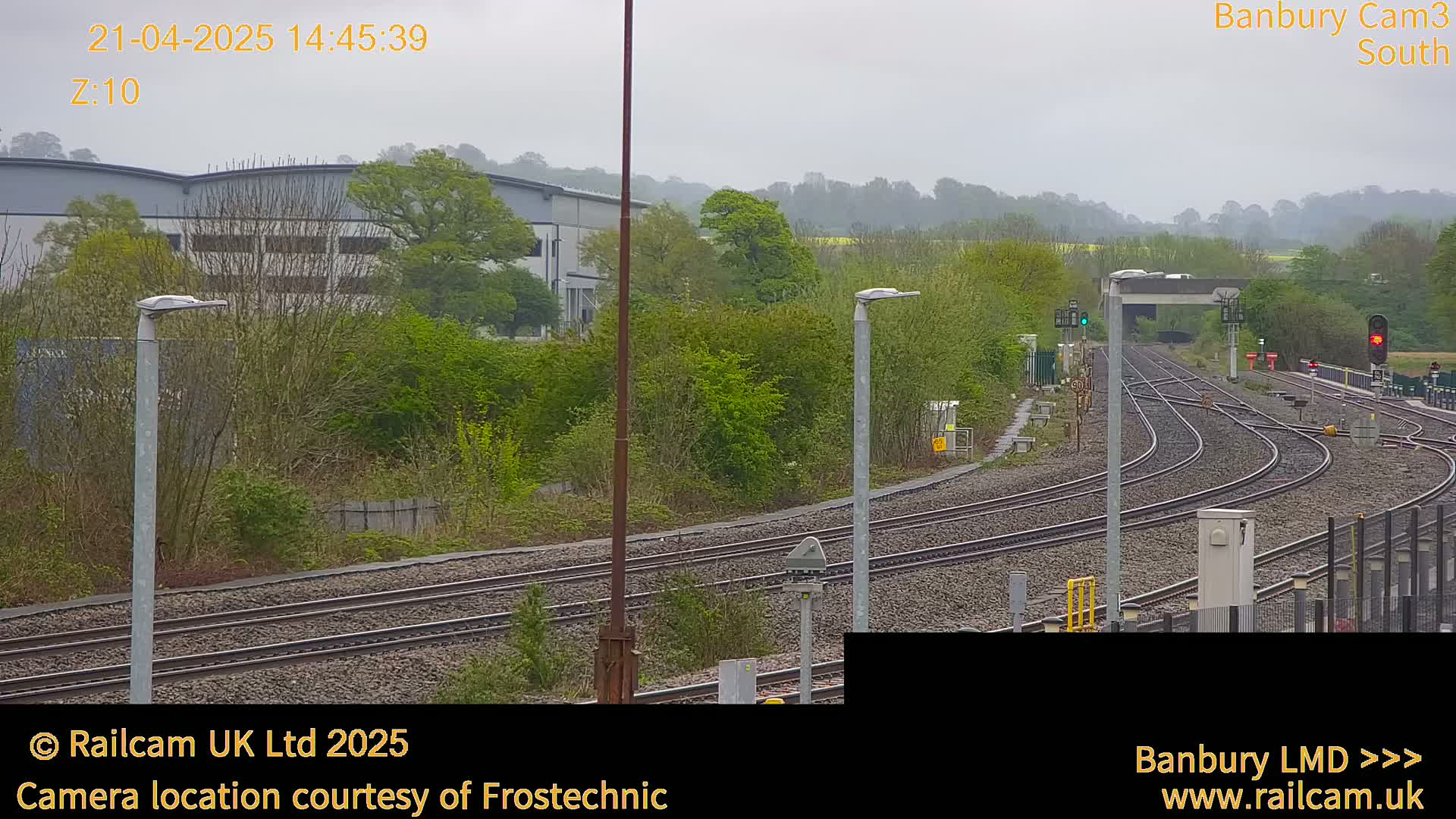 A view of train tracks curving through a landscape of green trees and a large gray building under an overcast sky.