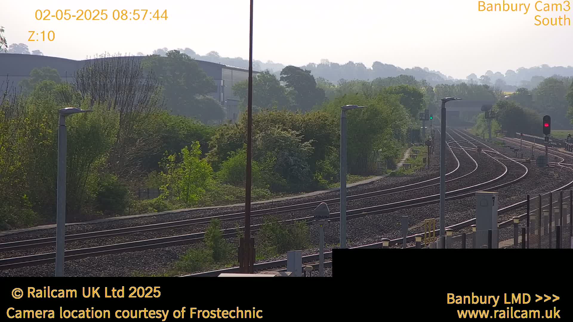 A hazy view of a train yard with multiple sets of tracks curving through a landscape of green trees and low-lying buildings under a bright, slightly hazy sky.