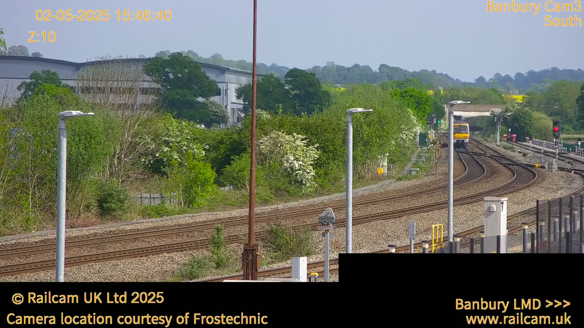 A yellow train is traveling along multiple train tracks on a sunny day, past lush green vegetation and industrial buildings.