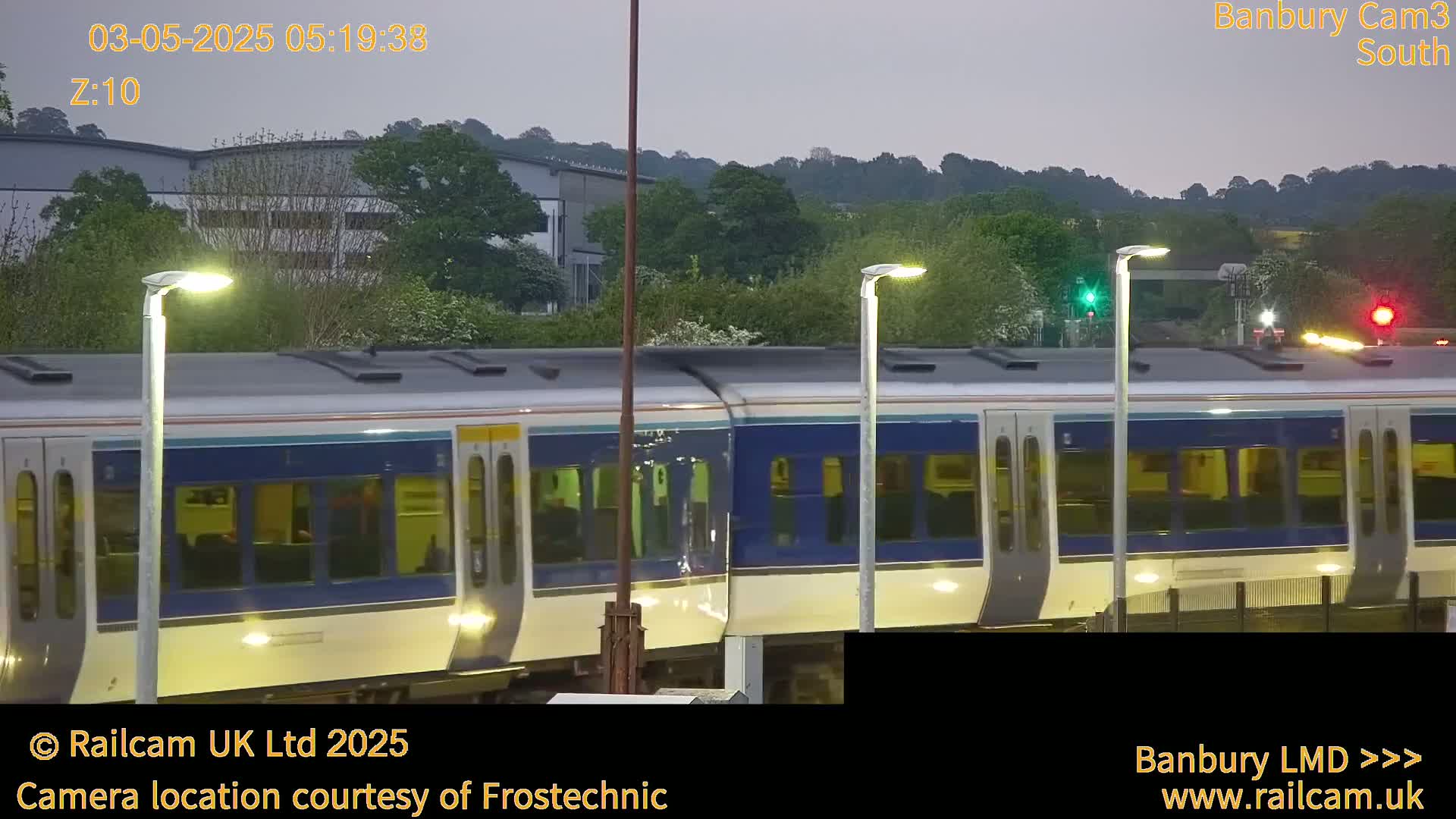 A train passes by under a twilight sky, beside lampposts and industrial buildings.